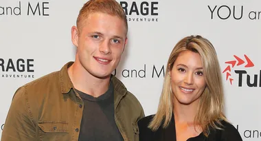 Two people smiling at an event, with promotional logos in the background.