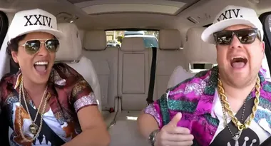 Two people wearing sunglasses and matching hats with "XXIV" inside a car, laughing and singing.