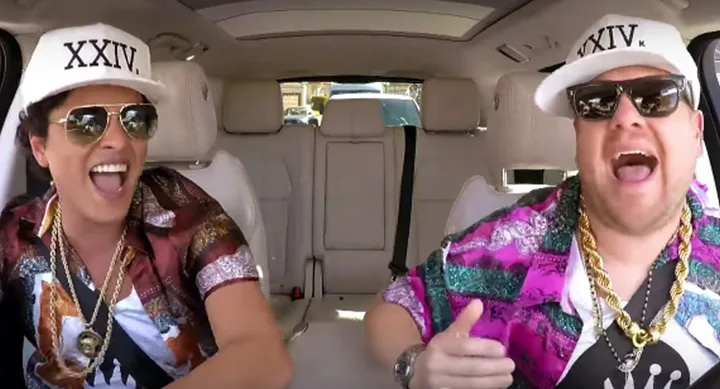 Two people wearing sunglasses and matching hats with "XXIV" inside a car, laughing and singing.