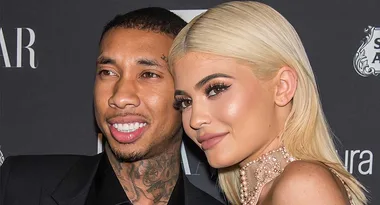 Two people smiling at an event with a dark background, one with blonde hair and the other with short hair and tattoos.