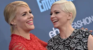 Two women smiling and embracing on a red carpet, one in a red lace dress, the other in a black and silver floral dress.