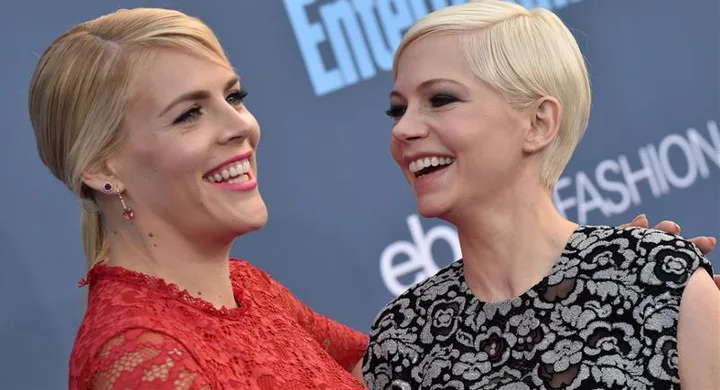 Two women smiling and embracing on a red carpet, one in a red lace dress, the other in a black and silver floral dress.