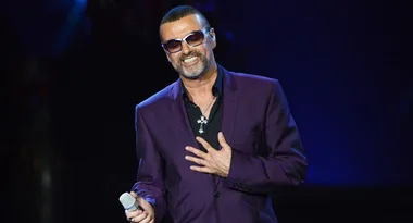 A man in a purple suit holds a microphone and smiles on stage.