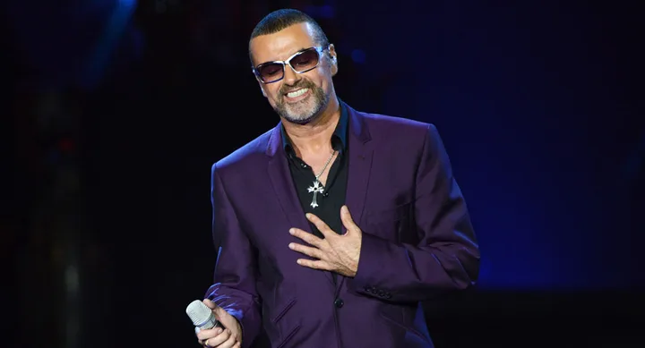 A man in a purple suit holds a microphone and smiles on stage.