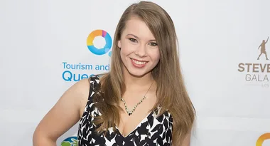 ```html
<img src="image.jpg" alt="Smiling woman in a black and white dress at an event, with logos in the background."/>
```
