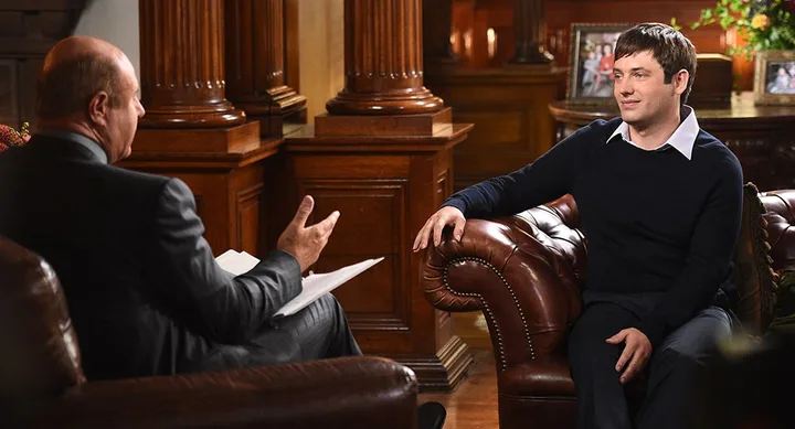Two men seated, having a conversation in a warmly lit room with wooden decor and framed photos in the background.