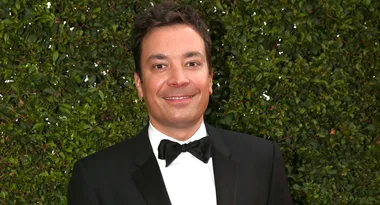 A man in a tuxedo with a bowtie stands in front of greenery, smiling at the camera.
