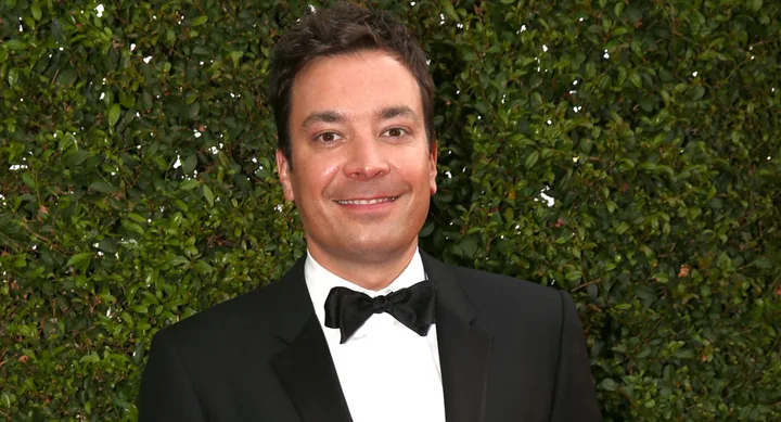 A man in a tuxedo with a bowtie stands in front of greenery, smiling at the camera.