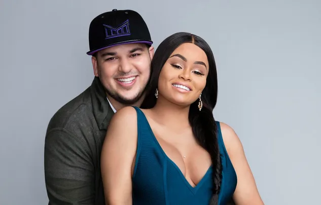 A smiling couple poses against a plain background; the man wears a hat, and the woman has long dark hair.