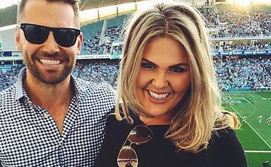 A man and woman smiling at a sports stadium with a crowd in the background.