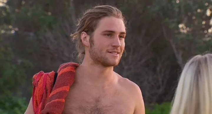 A man with long hair, holding a red towel, stands shirtless outside, talking to someone with long blonde hair.