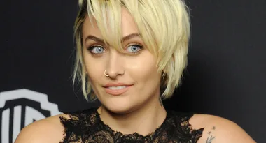 Blonde woman with short hair and black lace top at an event, smiling slightly against a dark backdrop.