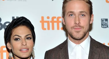 Two people pose in front of a TIFF backdrop, one wears a dark headwrap, the other a suit.