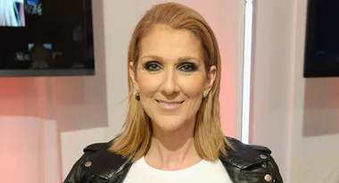 Adult with shoulder-length hair, wearing a black jacket, smiling at the camera in an indoor setting.