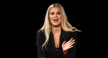 A woman with long blonde hair wearing a black outfit gestures on stage against a dark background.
