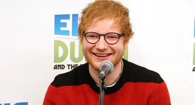 Man with red hair and glasses smiles at a microphone during an event; text behind partially visible.
