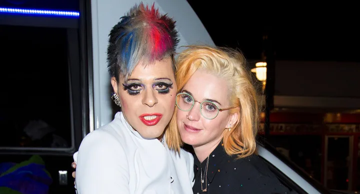 Two people posing together; one has colorful spiked hair and bold makeup, the other has glasses and blonde hair.