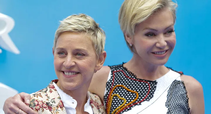 Two smiling individuals with short blonde hair, one in a floral jacket and the other in a patterned dress, against a blue background.