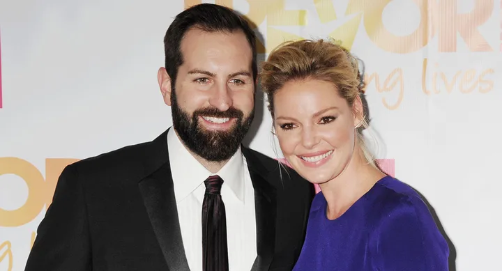 Two people smiling at a red carpet event, one in a blue dress and the other in a black suit.