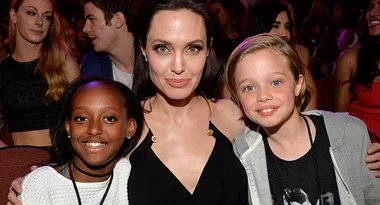 A woman in a black dress is seated with two children, one on each side, surrounded by people in an event setting.