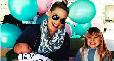 A woman with sunglasses smiles, holding a baby, next to a child; colorful balloons in the background.