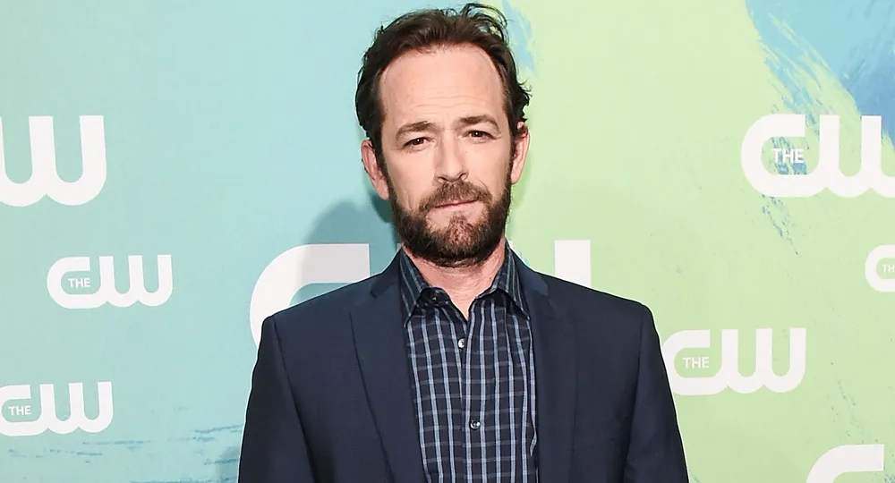 Man in a dark suit and checkered shirt stands against a green CW backdrop.
