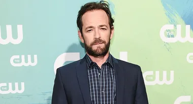 Man in a dark suit and checkered shirt stands against a green CW backdrop.