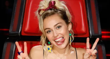 ```plaintext
Person with blonde hair making peace signs and smiling in a red chair.
```