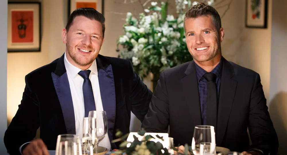 Two men in suits smiling at a table, with dinner settings and floral decorations in the background.