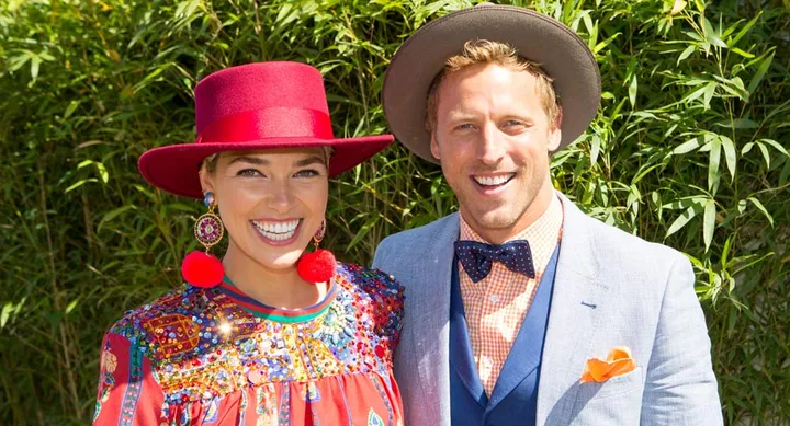 Two people smiling outdoors; woman in colorful dress and red hat, man in light suit and hat, both in front of green foliage.