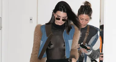 Two women walking indoors, one wearing sunglasses and a sheer top with a denim and fur jacket.