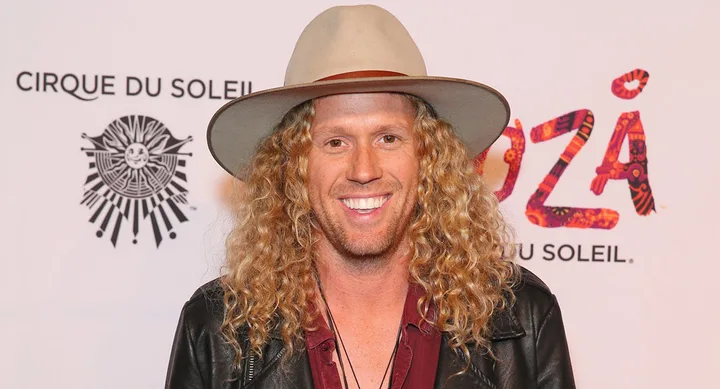 Smiling person with curly hair and hat at Cirque du Soleil event backdrop.