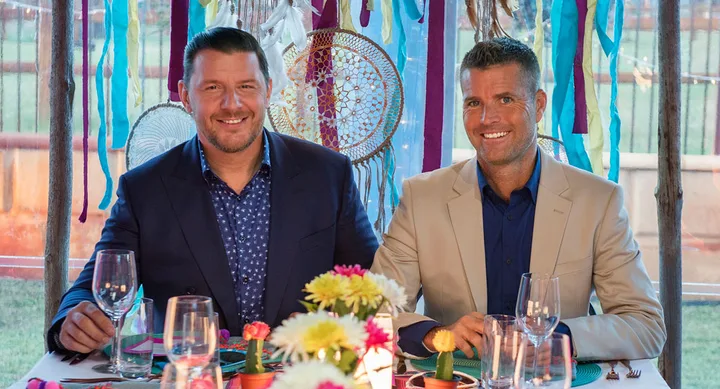 Two men in suits sitting at a colorful table with dreamcatchers and ribbons in the background.