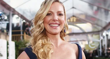 A woman with wavy blonde hair in a formal dress smiles broadly on a red carpet.