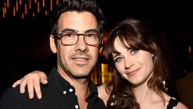 A man with glasses and a woman smiling, both looking at the camera in a dimly lit setting.