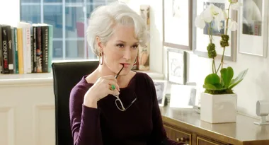 Character with short white hair in an office, holding glasses to lips, next to books and a plant.