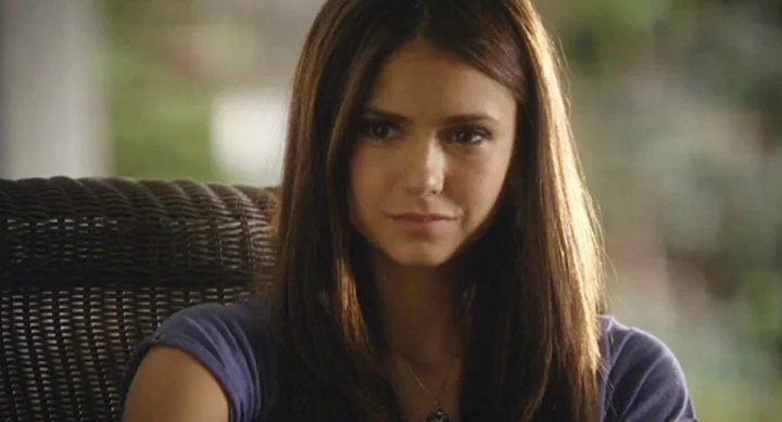 ```plaintext
Woman with long brown hair sits on a wicker chair, wearing a blue top, looking forward.
```