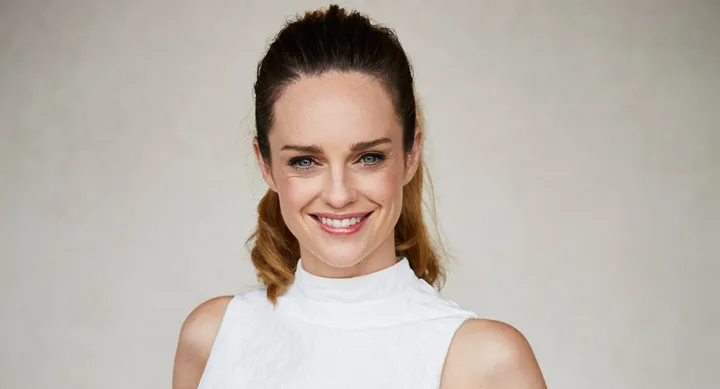 A woman with long hair smiles, wearing a sleeveless white top against a neutral background.