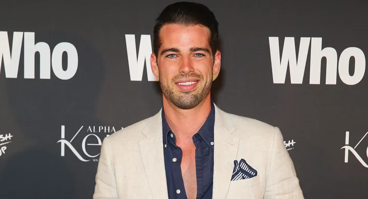 A man in a beige suit and blue shirt smiles on the red carpet with "Who" in the background.