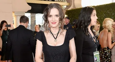 A woman in a black dress poses on a red carpet, smiling, with other attendees and media in the background.