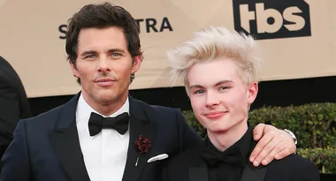 Two people in formal suits pose together on a red carpet at an event.