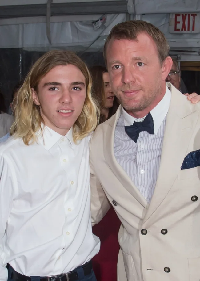 Two men in dress shirts and blazers pose together at a formal event. One has long blonde hair, the other short brown hair.
