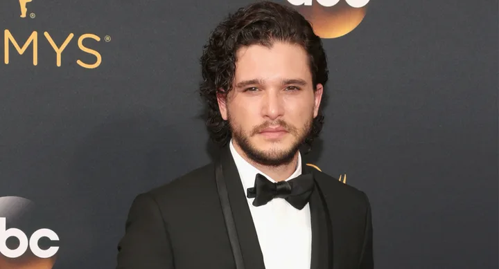 Actor in a black tuxedo at an awards event, standing in front of an "Emmys" backdrop.