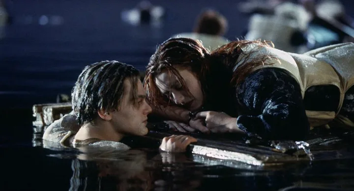 Two characters from "Titanic" in the ocean; one clings to a door, while the other looks up at them, both wearing life vests.