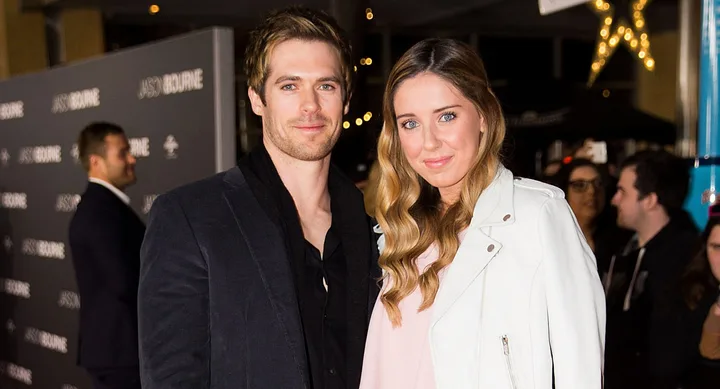 A man in a black jacket and a woman in a white jacket smiling at a "Jason Bourne" event with a crowd in the background.