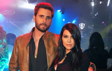 A man in a snakeskin jacket and a woman in black stand together in a dimly lit club with colorful lights.