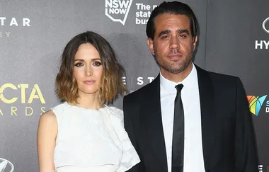 A man and a woman pose at the AACTA Awards, standing in front of a banner with event logos.