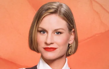 A woman with short blond hair and earrings smiles against an orange background.