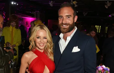 Kylie Minogue in a red dress with a bearded man in a suit at an event, surrounded by people in a dimly lit room.