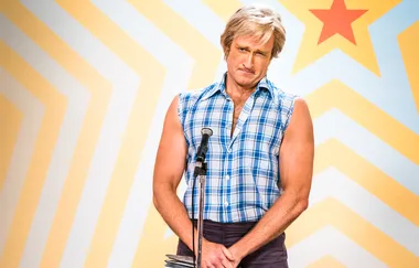 A man in a sleeveless plaid shirt stands at a microphone with a retro star background.